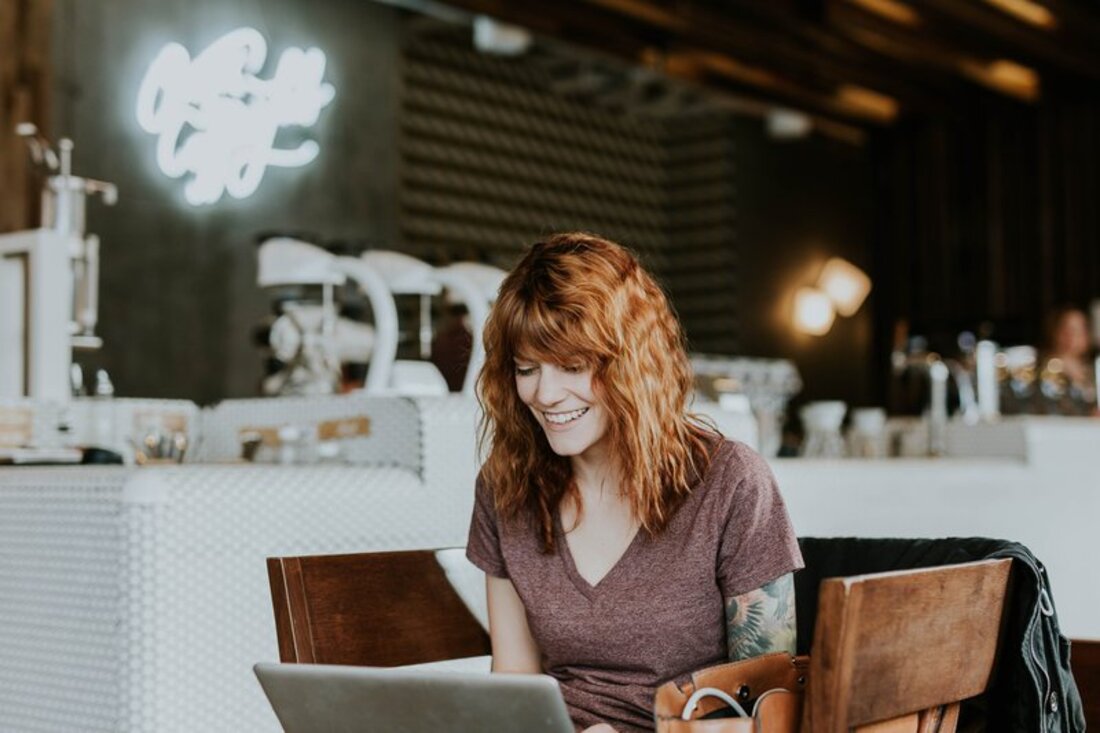 Image contains a woman working on her laptop and smiling, sitting in a coffee shop