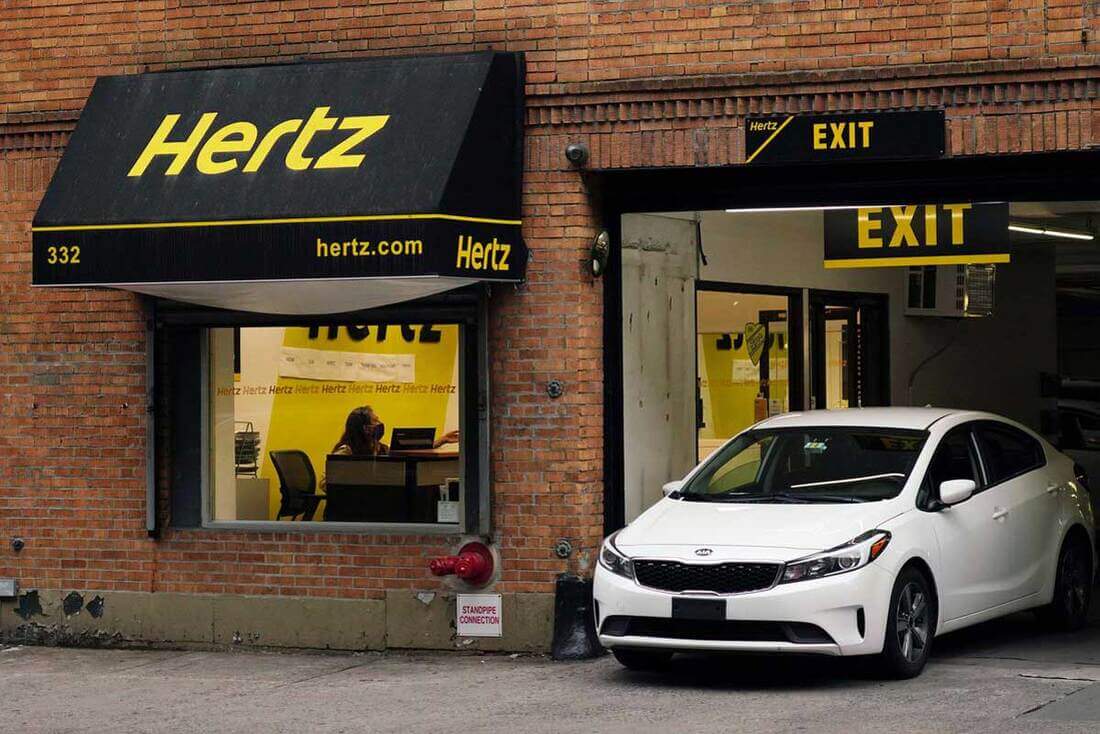 Image for why did hertz go bankrupt? A Hertz dealership in a brick and mortar building, with a white vehicle exiting on the right 