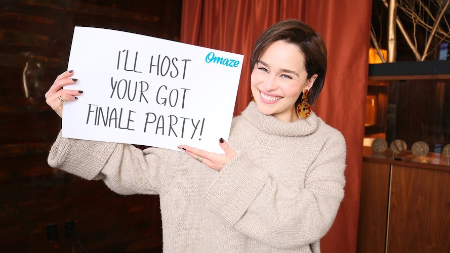 Image for Is Omaze a Scam: Emilia clark is holding a sign that invites others to let her host a finale party, with a studio in the background